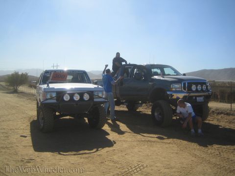 1990 Ford Bronco - white SUV and a gray lifted Ford pickup with roof lights are parked on dirt as several people gather around.
