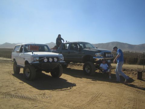 1990 Ford Bronco - white lifted SUV and a gray Ford pickup with roof lights sit parked on dirt while several people stand nearby.