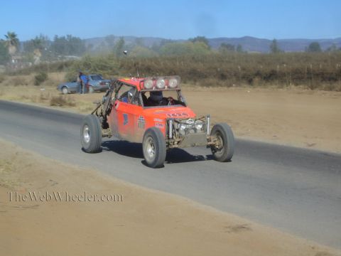 1990 Ford Bronco - orange off-road race buggy with three roof lights speeds along a paved road past a parked sedan.