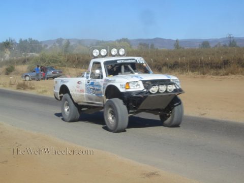 1990 Ford Bronco - white Ford off-road race truck with a roof light bar and tube bumper speeds along a paved desert road.
