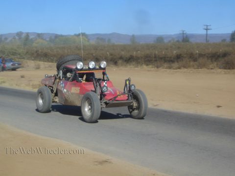1990 Ford Bronco - dark red off-road race buggy carrying a spare tire and roof lights races along a paved desert road.