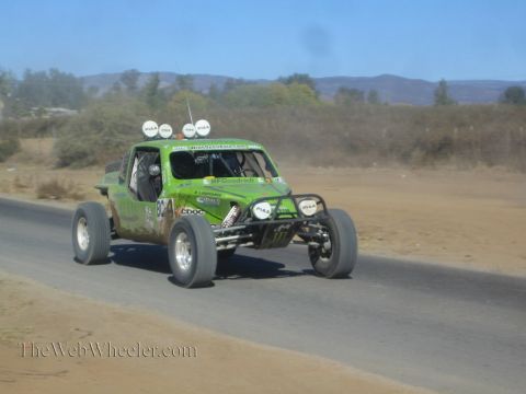 1990 Ford Bronco - bright green off-road race buggy with roof lights speeds along a paved road through dry hill country.