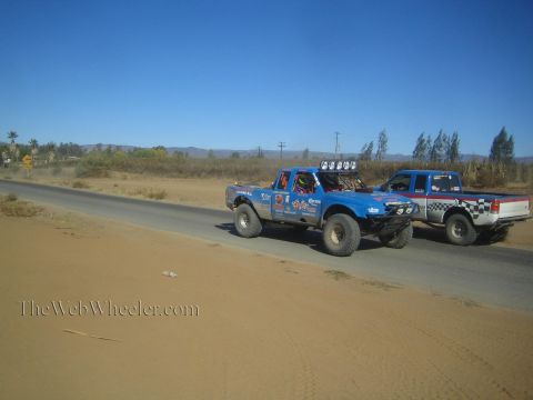 1990 Ford Bronco - blue Ford off-road race truck with roof lights and a silver pickup travel along a paved desert road.