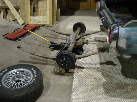 1992 Ford Crown Victoria - Rear axle assembly and leaf springs are laid out on the floor next to a tire and a jack, suggesting a vehicle repair or restoration project.