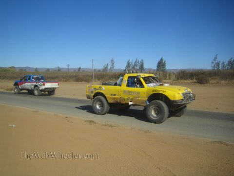 1990 Ford Bronco - yellow off-road race truck with a roof light bar and a blue pickup travel along a paved desert road.