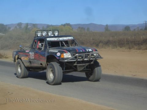 1990 Ford Bronco - black off-road race truck numbered 718 in Herrera's Motorsports livery speeds along a paved desert road.
