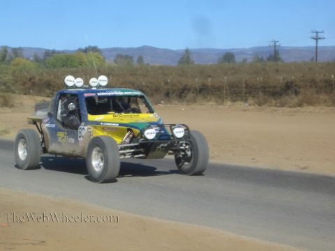 1990 Ford Bronco - blue and yellow off-road race buggy with four roof lights races along a paved road in dry hill country.