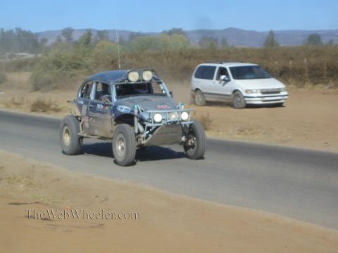 1990 Ford Bronco - blue Volkswagen Beetle-bodied off-road race buggy catches air on a paved desert road as a white minivan follows behind.