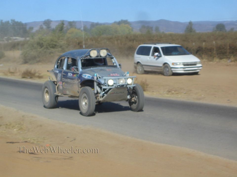 Blue VW Beetle-based off-road race buggy mid-jump on a paved road, white minivan trailing