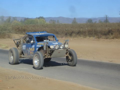 1990 Ford Bronco - blue open tube-frame off-road race buggy marked 517 speeds along a paved road through dry desert hills.