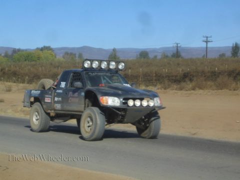 1990 Ford Bronco - black off-road race truck numbered 706 with a roof-mounted light bar drives along a paved road in dry hills.