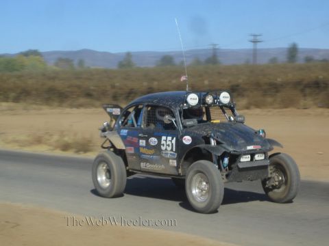1990 Ford Bronco - black off-road race buggy marked 551 with roof light pods races along a paved road in dusty desert terrain.