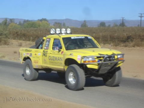 1990 Ford Bronco - yellow off-road race truck lettered Guzman Construction and numbered 740 catches air on a paved desert road.