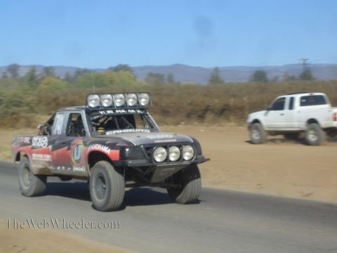 1990 Ford Bronco - black and red off-road race truck with a five-light roof bar speeds down a paved road as a white pickup trails in the dust.