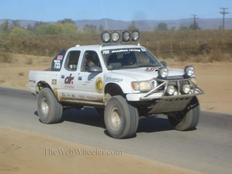 1990 Ford Bronco - white Toyota double-cab off-road race truck numbered 755 races along a paved road through dry desert terrain.