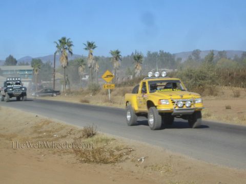 1990 Ford Bronco - yellow off-road race truck speeds along a paved road past palm trees and a roadside town, with another racer behind.