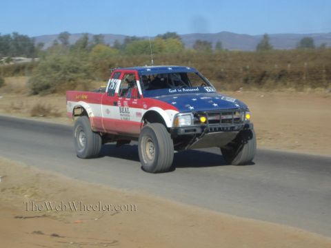 1990 Ford Bronco - patriotic stars-and-stripes painted off-road race truck catches air on a paved road through dusty desert hills.