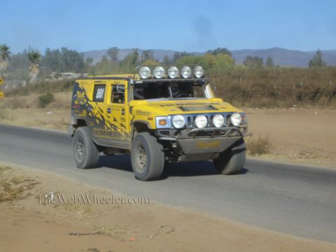 1990 Ford Bronco - yellow Hummer-style off-road race vehicle numbered 861 with a roof light bar drives along a paved desert road.