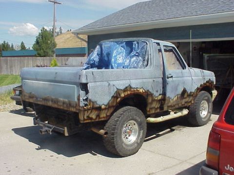 1989 Ford Bronco - Passenger-side rear three-quarter view of a topless full-size Ford Bronco in a driveway, with the hardtop removed and a blue tarp bunched o…