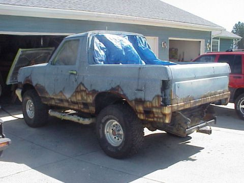1989 Ford Bronco - Driver-side rear three-quarter view of a gray full-size Ford Bronco with the hardtop removed and a blue tarp draped over the interior, park…