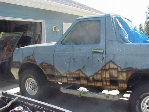 1989 Ford Bronco - Passenger-side view of a full-size Ford Bronco in mid-paint, its blue-gray body wearing a freshly airbrushed mountain-range mural along the…