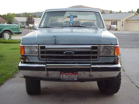1989 Ford Bronco - Head-on front view of a faded blue-gray Ford Bronco parked on a residential driveway, showing the bar-style grille with blue Ford oval, chr…