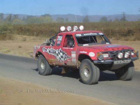 1990 Ford Bronco - red off-road race truck numbered 758 with checkered-flag graphics speeds along a paved road through dry hills.