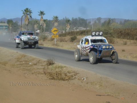 1990 Ford Bronco - Two off-road race buggies, one with stars-and-stripes graphics, run a paved road past palm trees and a roadside town.