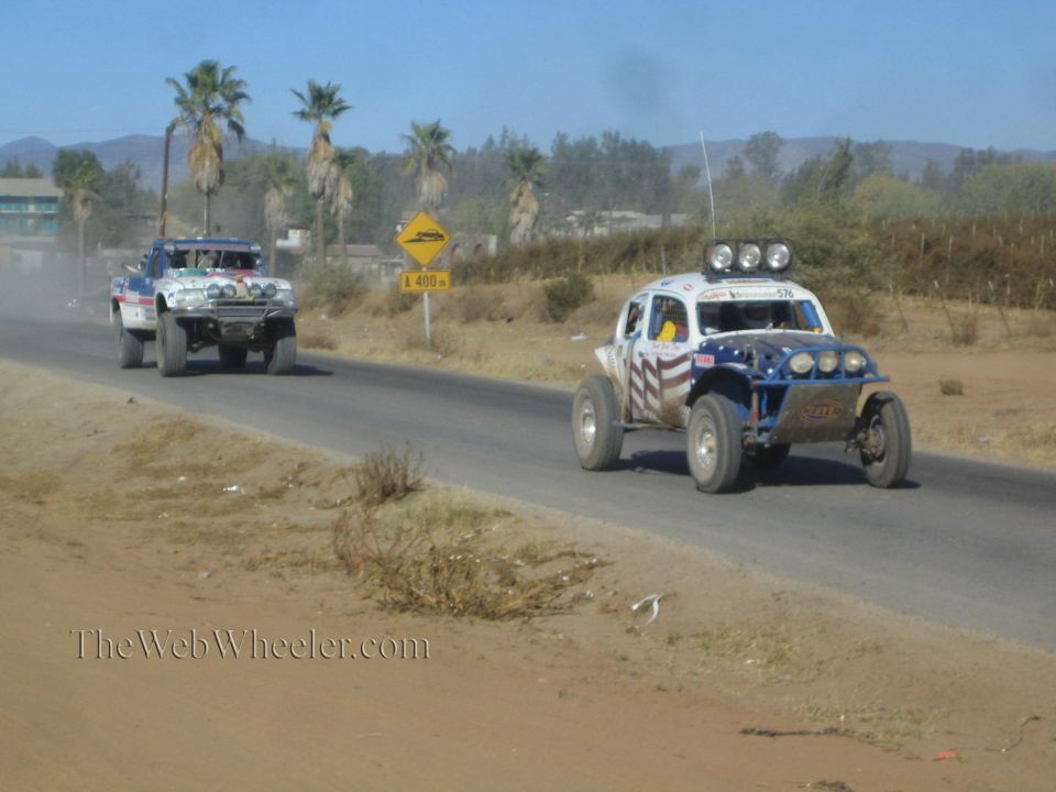 Two off-road race buggies on a paved road past palm trees, one in stars-and-stripes