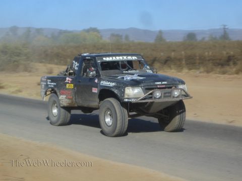 1990 Ford Bronco - black off-road race truck with a roof light bar speeds along a paved road through dry, dusty desert terrain.