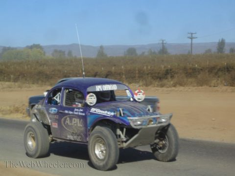 1990 Ford Bronco - purple Volkswagen Beetle-based race buggy lettered A.P.V. drives along a paved road through dry desert terrain.