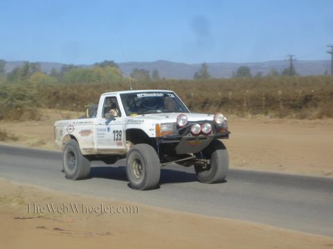 1990 Ford Bronco - white off-road race pickup numbered 739 catches air on a paved road through dry desert hills.