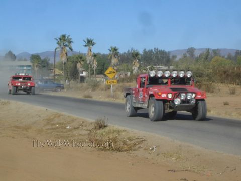 1990 Ford Bronco - red Hummer-style off-road race vehicle with a roof light bar runs a paved road past palm trees, another racer ahead.