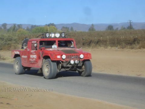 1990 Ford Bronco - red Hummer-style off-road race vehicle numbered 803 with a roof light bar drives along a paved desert road.