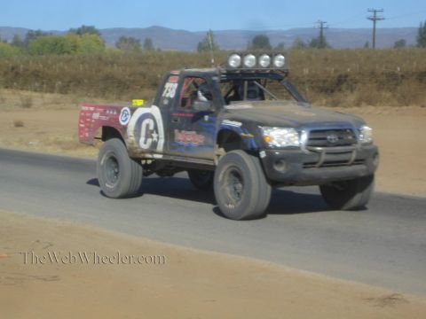 1990 Ford Bronco - black Toyota off-road race truck numbered 735 with a roof light bar speeds along a paved road in dry desert terrain.