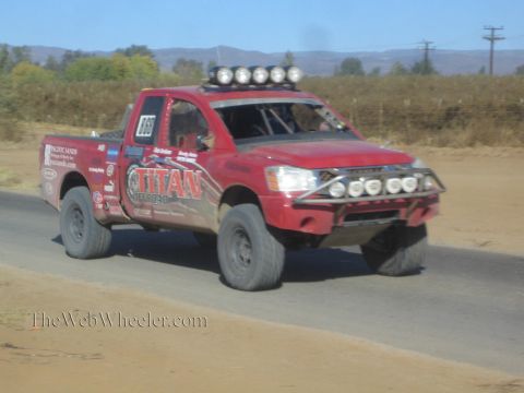 1990 Ford Bronco - red Nissan Titan off-road race truck with a roof light bar drives along a paved road through dry desert terrain.