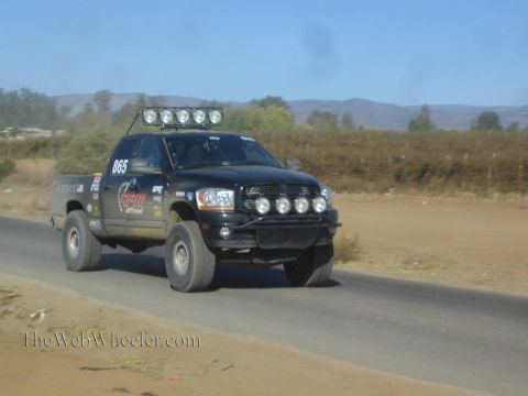 1990 Ford Bronco - black Dodge Ram off-road race truck numbered 865 with a roof-mounted light bar speeds along a paved desert road.