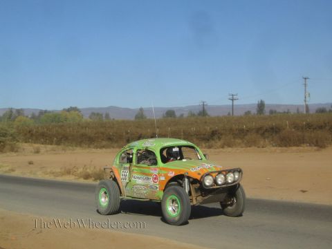 1990 Ford Bronco - green and orange Volkswagen Beetle-bodied off-road race car with front light pods drives along a rural paved road.