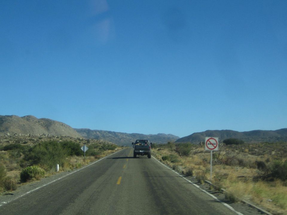View through windshield of desert highway with a dark truck ahead and a no-passing sign