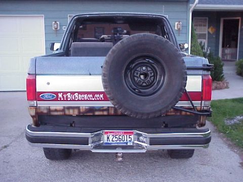 1989 Ford Bronco - Straight-on rear view of a two-tone dark blue and silver Ford Bronco with the hardtop removed, a full-size spare mounted on a swing-out tir…