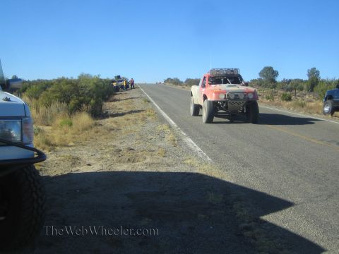 1990 Ford Bronco - orange off-road race truck travels a rural highway near parked support trucks and roadside spectators.