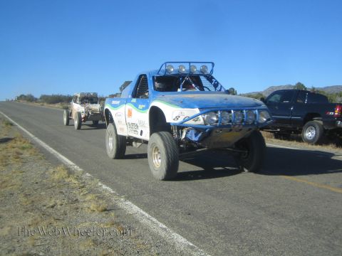 1990 Ford Bronco - blue and white off-road race truck with a roof light bar travels a rural highway, with another buggy and an SUV nearby.