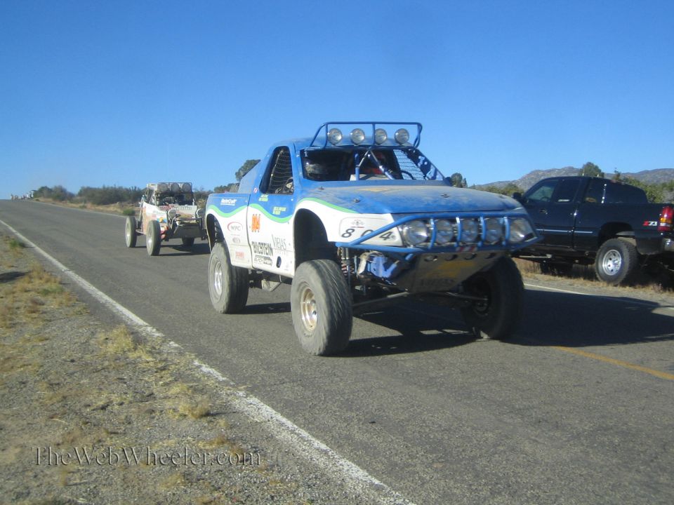 Blue and white off-road race truck towing a buggy on a rural highway