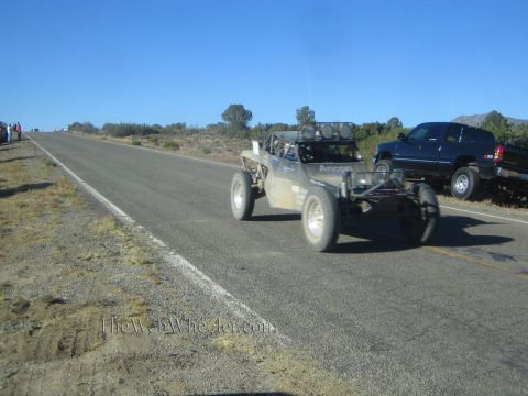1990 Ford Bronco - tan tube-frame single-seat off-road race buggy drives along a rural road next to a parked dark SUV.