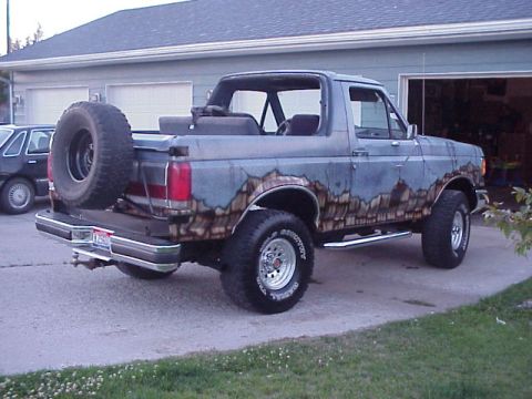 1989 Ford Bronco - 3/4 rear passenger-side view of a topless full-size Ford Bronco in a driveway, painted light blue with a rock/mountain-scene mural along th…