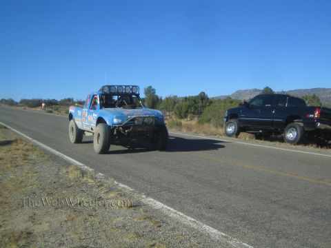 1990 Ford Bronco - blue off-road race truck with roof lights travels a rural highway as a dark pickup sits parked on the shoulder.