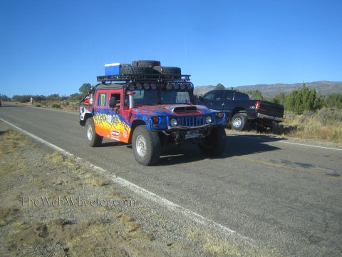 1990 Ford Bronco - brightly flame-painted Hummer H1 with a roof rack, spare tire and light bar drives a rural highway past a parked pickup.
