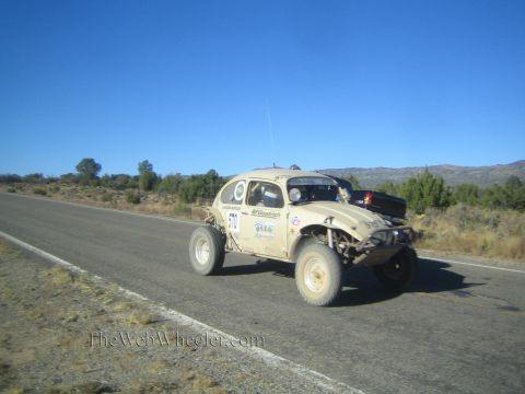1990 Ford Bronco - tan Volkswagen Beetle-bodied off-road race car drives a rural highway with hills and trees in the background.