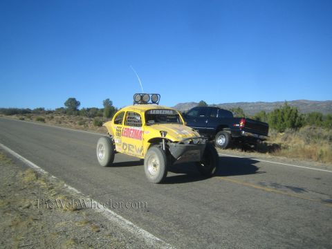 1990 Ford Bronco - yellow Volkswagen Beetle off-road race car marked Ledesma 555 drives a rural highway past a parked dark pickup.