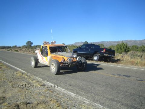 1990 Ford Bronco - orange and white tube-frame off-road race buggy drives a rural highway as a dark pickup sits on the shoulder.